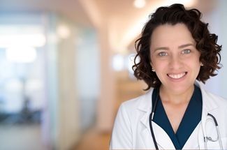 Simling woman with short, curly dark brown hair wearing a white doctor's coat with a light pink background